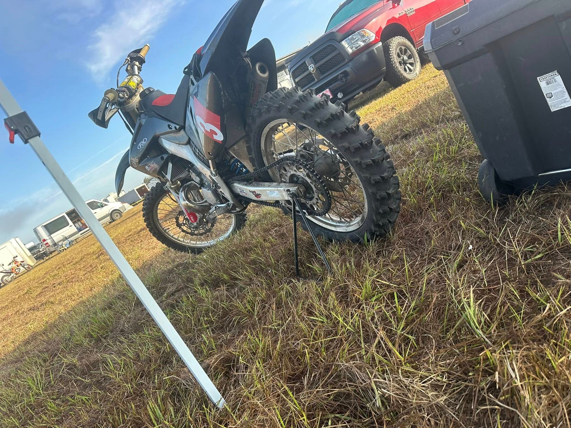 A black and red dirt bike parked on grass near a red truck and a grey storage bin under a clear sky.