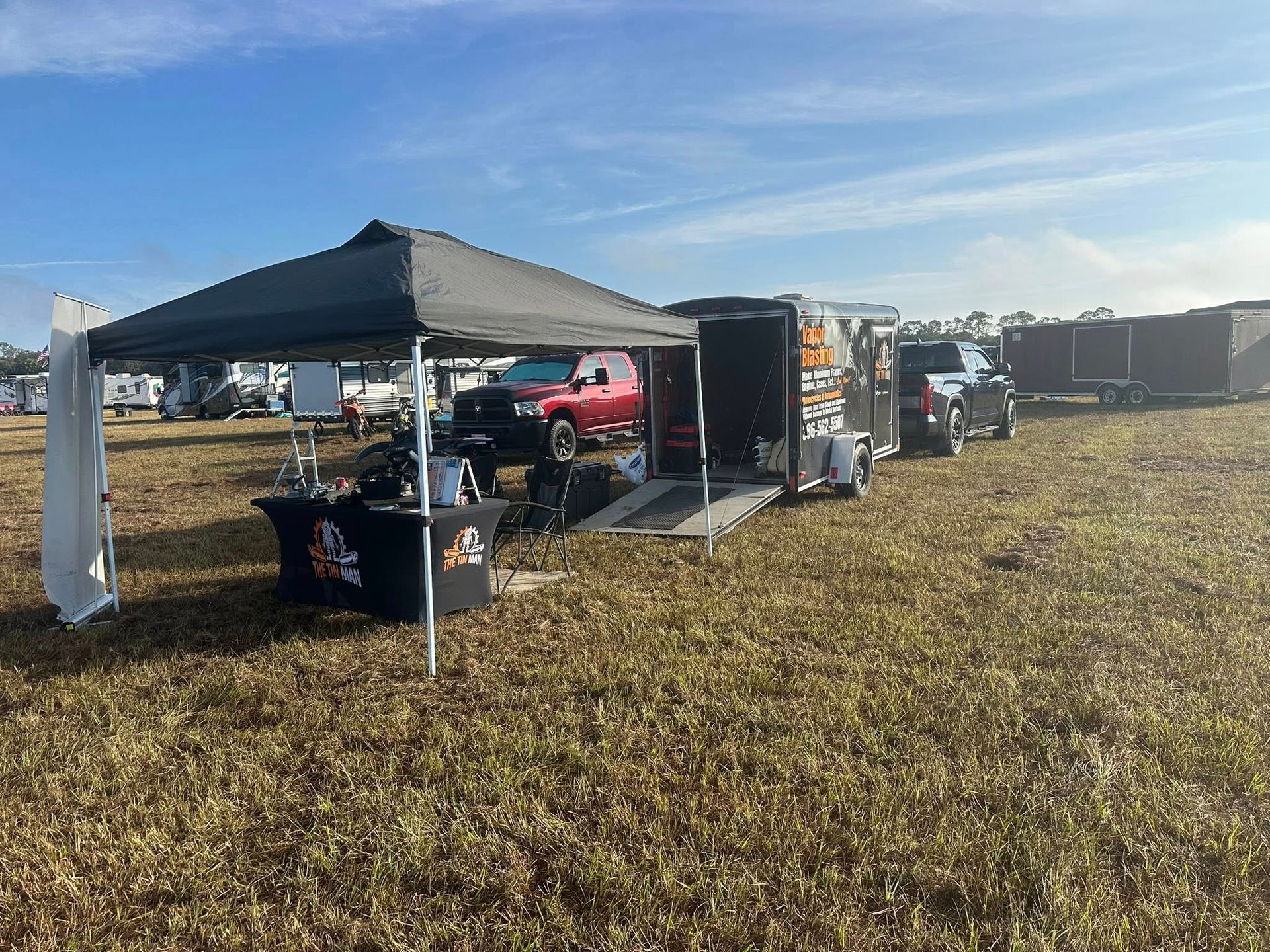 A black canopy tent with a branded table sits in a field next to an open equipment trailer and a red pickup truck.