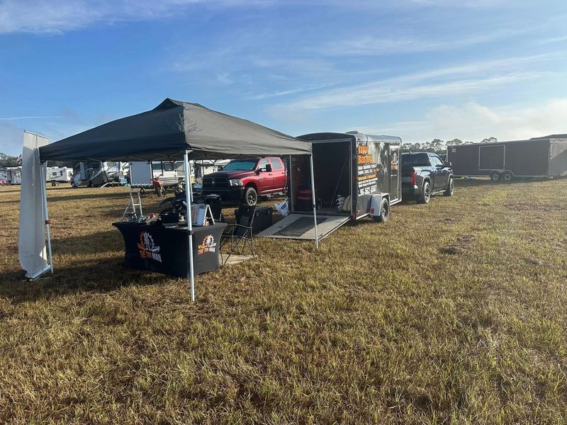 A black canopy tent with a branded table sits in a field next to an open equipment trailer and a red pickup truck.