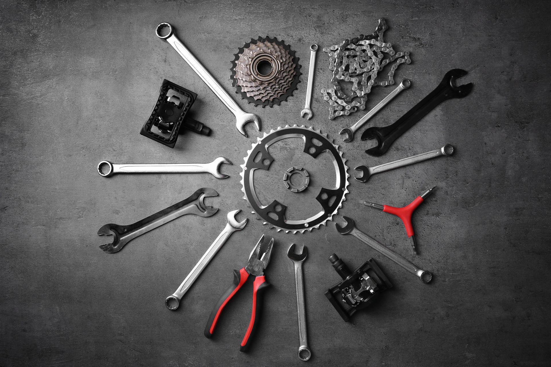 Bicycle repair tools and parts, including wrenches, pedals, a chain, and gears, arranged in a circle on a gray surface.