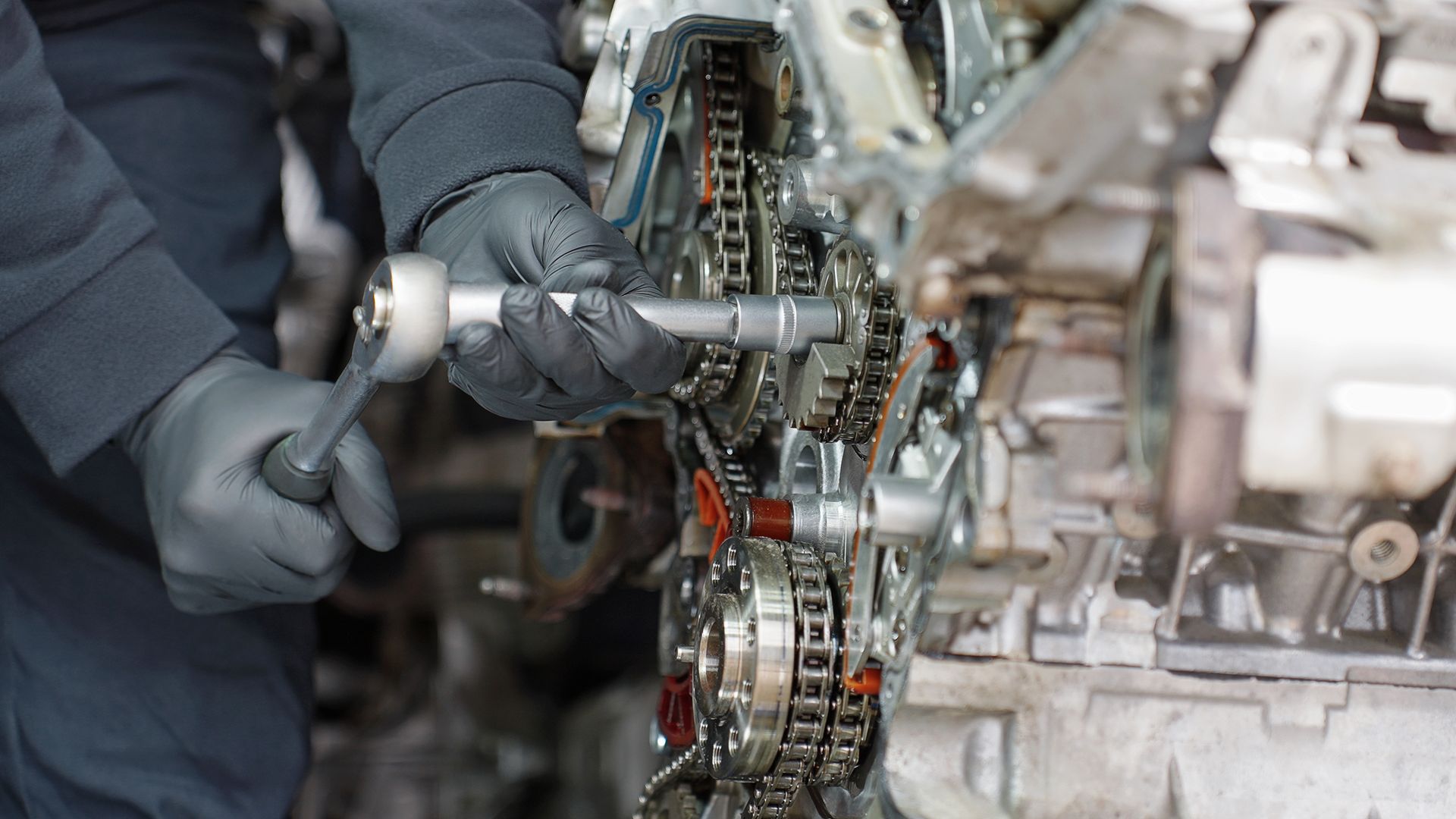 A mechanic wearing black gloves uses a ratchet tool to work on the internal timing chain mechanism of an engine.
