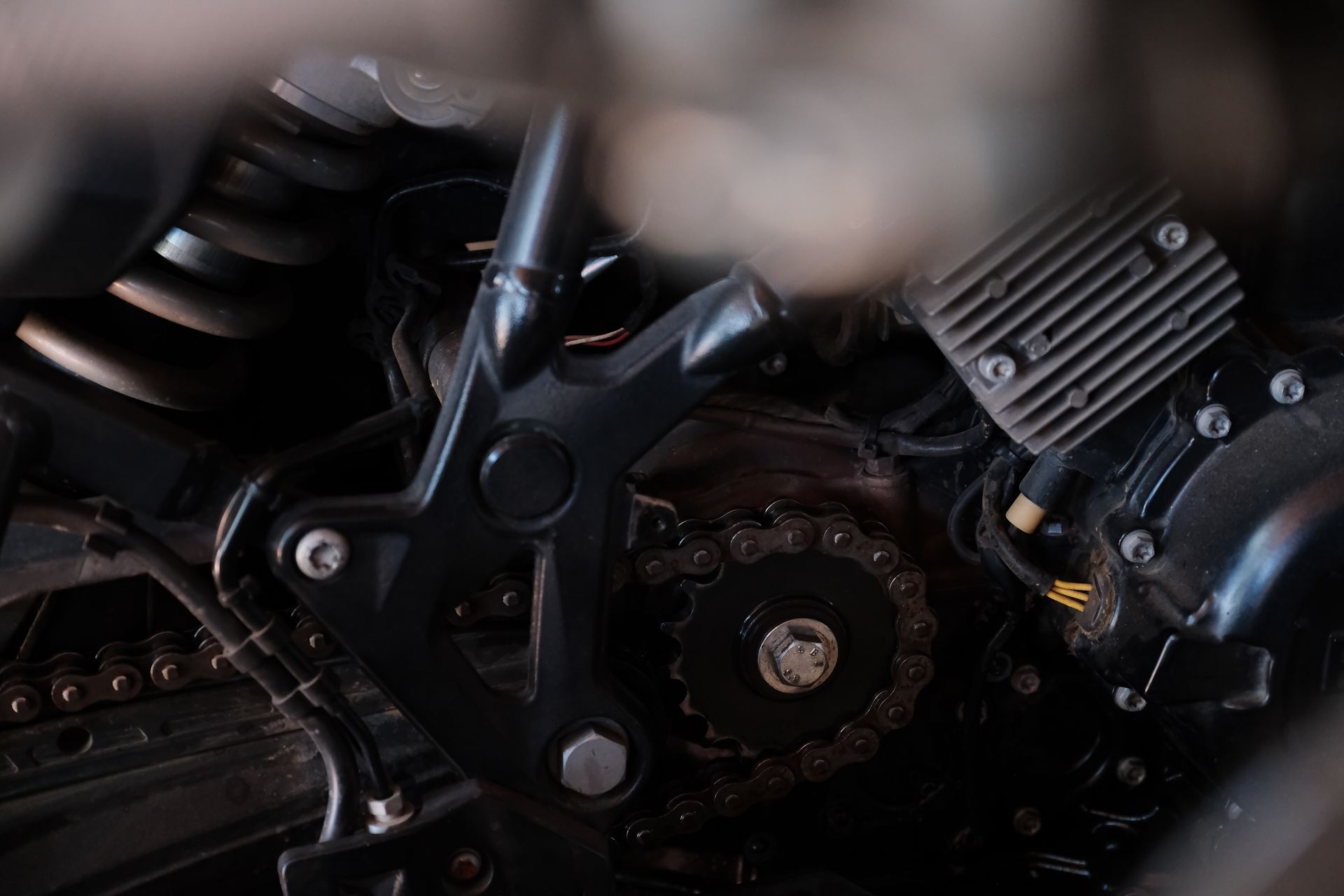 Close-up of a motorcycle's mechanical drivetrain, showing the drive sprocket, chain, and frame structure in dark tones.