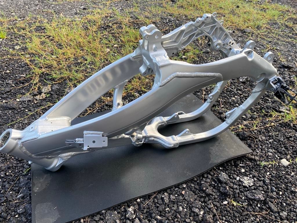 A silver aluminum motorcycle frame sitting on a black mat outdoors on a gravel surface.