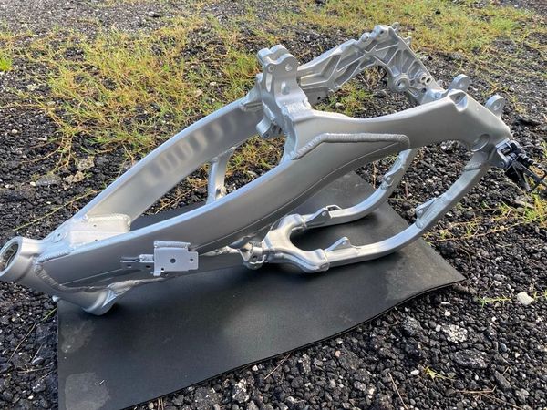 A silver aluminum motorcycle frame sitting on a black mat outdoors on a gravel surface.