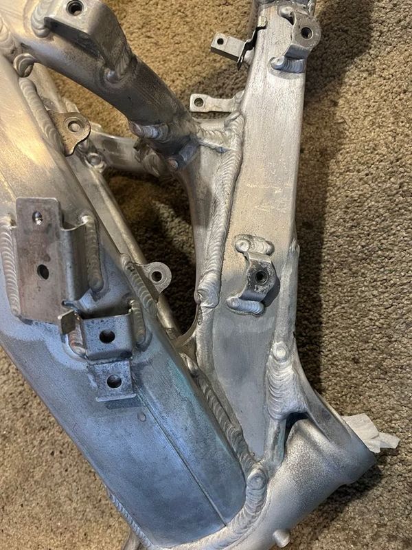 A close-up view of an unfinished aluminum motorcycle frame showing various welded mounting brackets and joints.