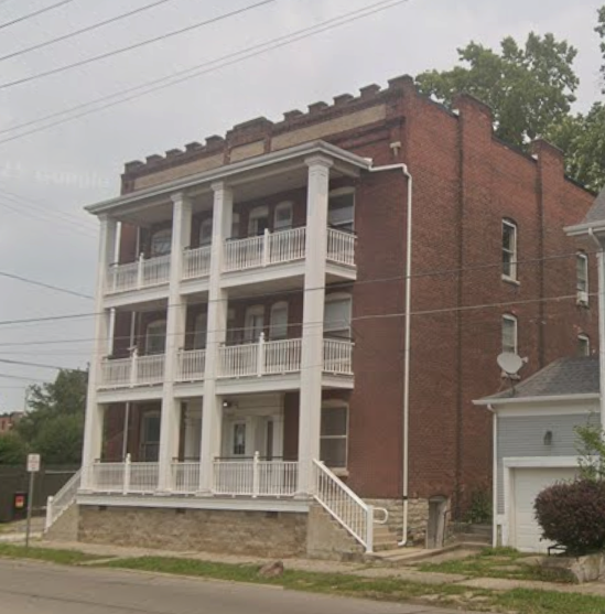 Three-story brick apartment building with white balconies and trim, on a street with a cloudy sky.