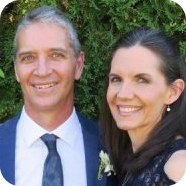 Man and woman smiling, posed closely together outdoors. Man in suit and tie. Woman in dark dress.