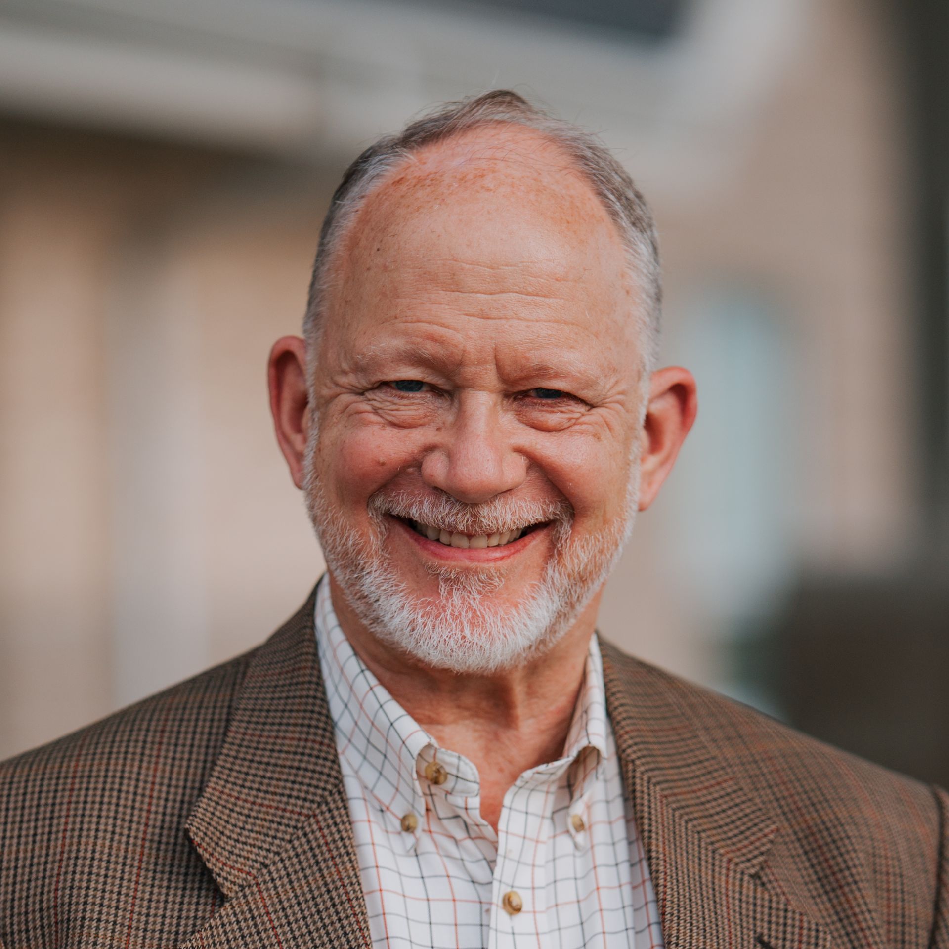 Smiling man with graying beard and brown blazer; outdoors.