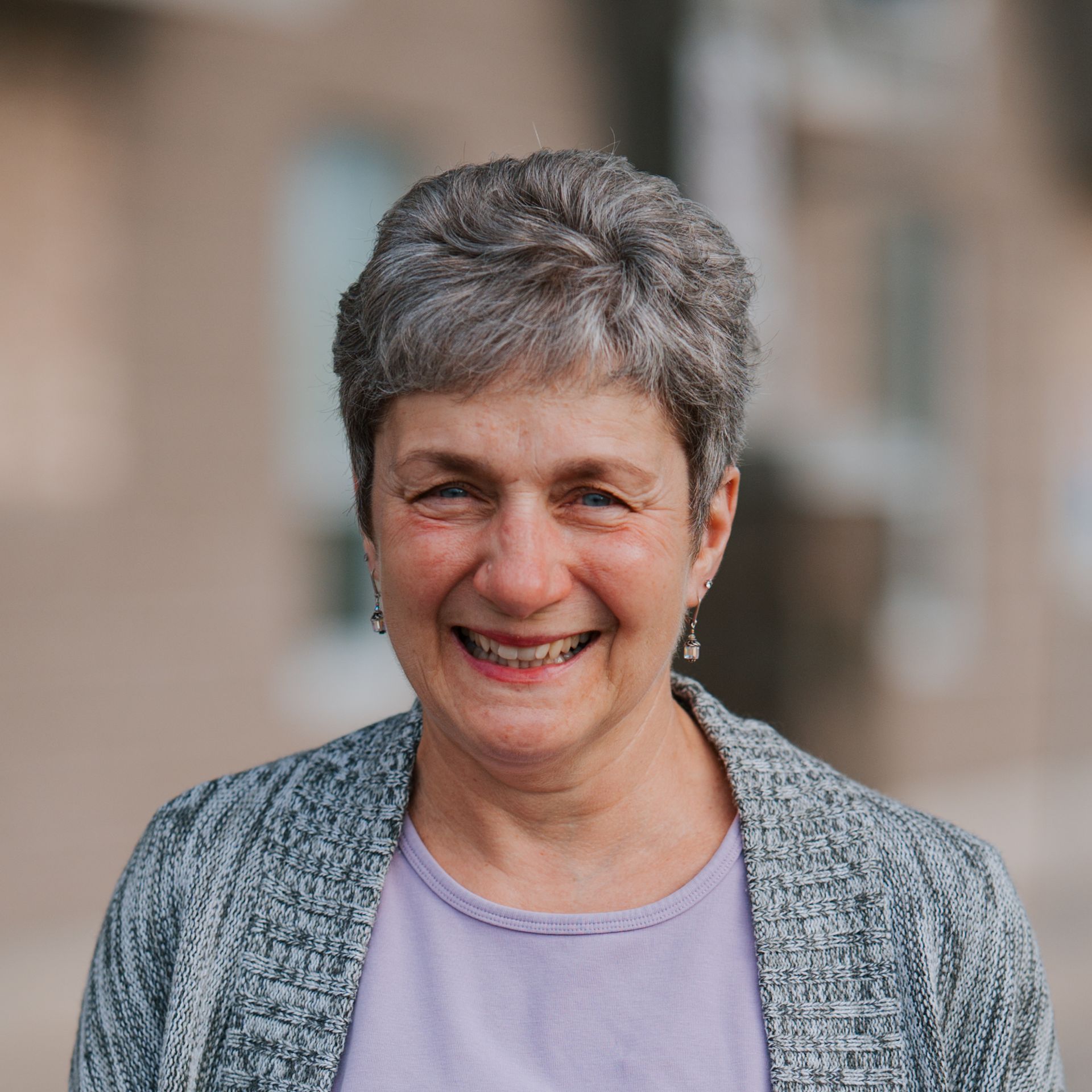 Woman with short gray hair smiles outdoors, wearing a gray cardigan and purple shirt.