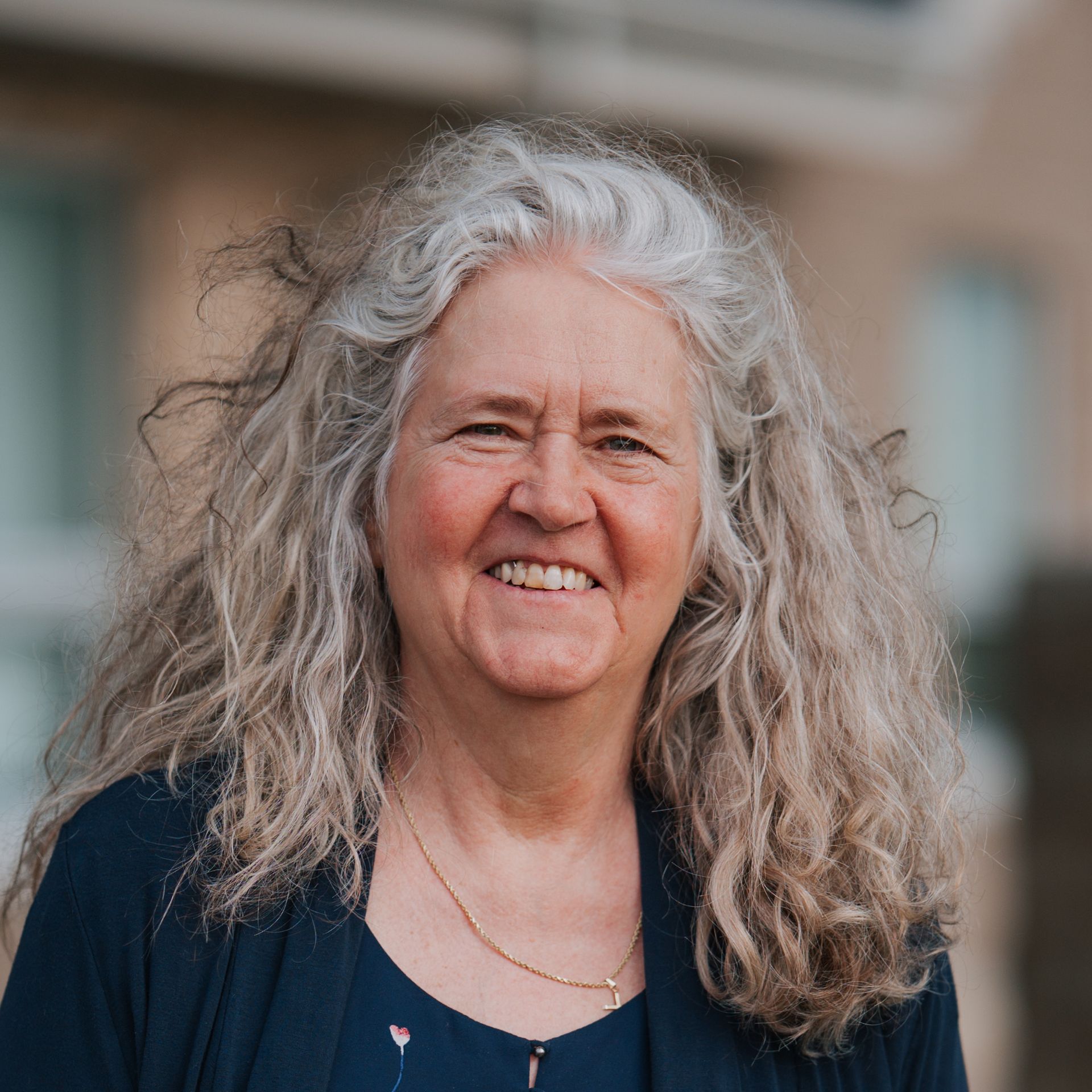 Woman with long, gray, curly hair smiling, wearing a dark blue top and necklace.