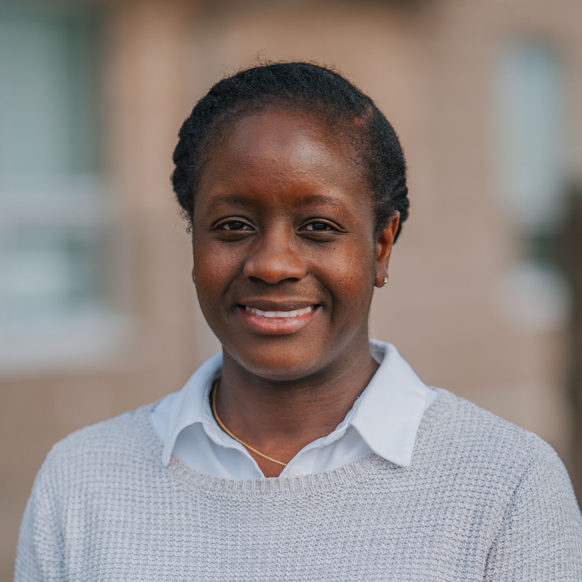 Smiling person in a light gray sweater and collared shirt outside a building.