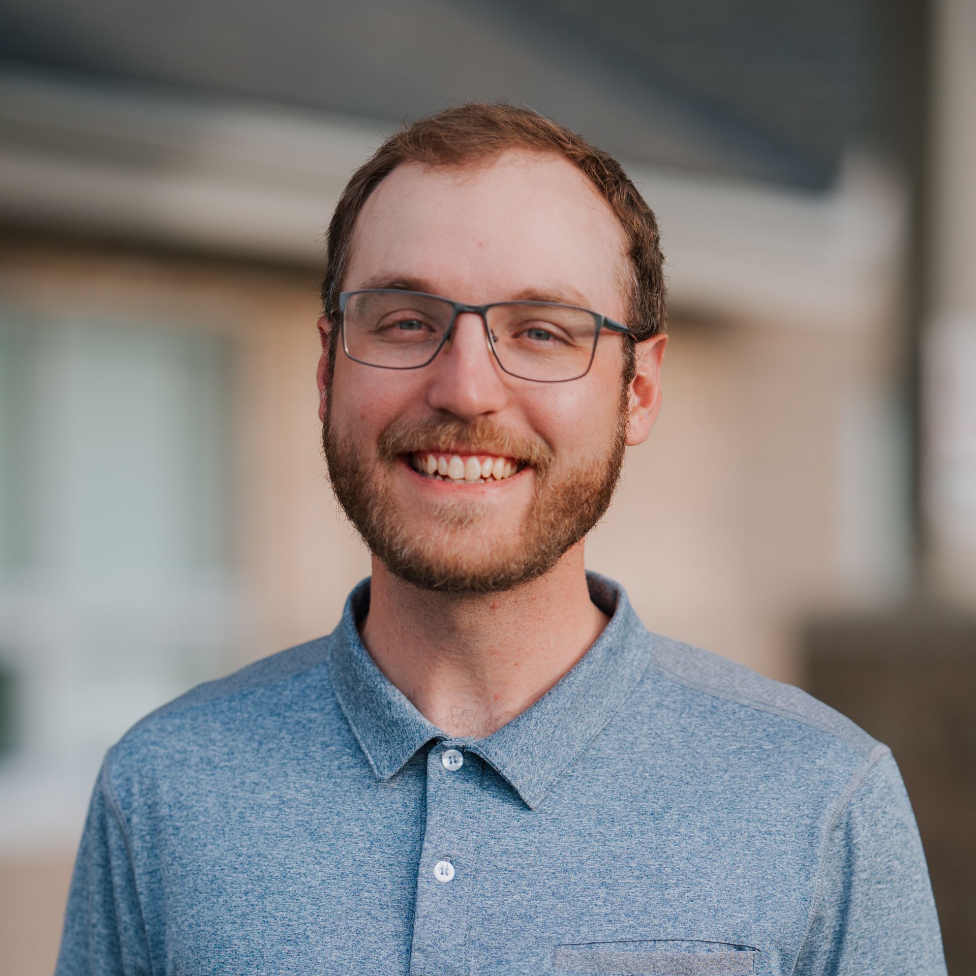 Man wearing glasses, smiling, with a beard, in a blue polo shirt, outdoors.
