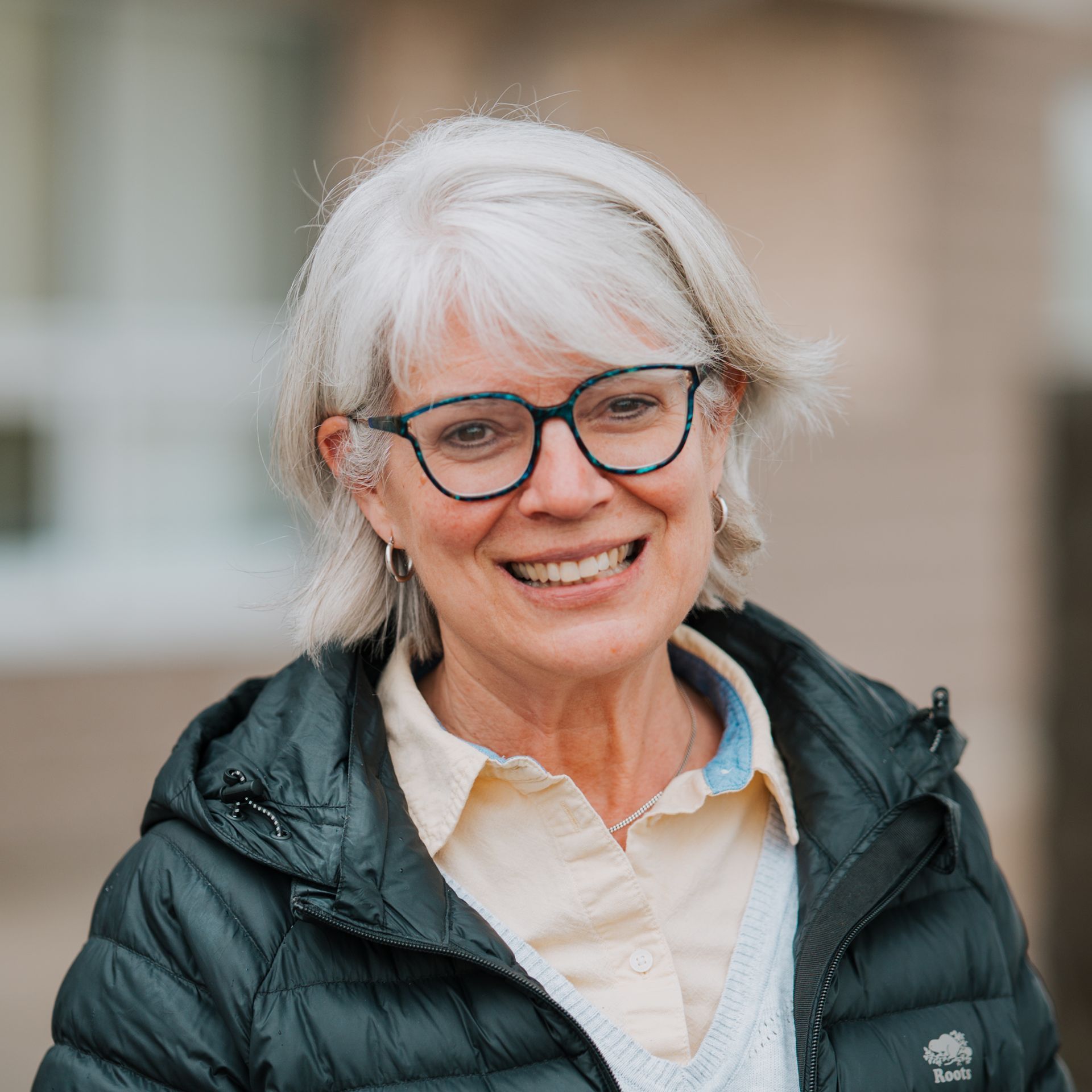 Woman with glasses smiles, wearing a black jacket and light yellow shirt, outdoors.