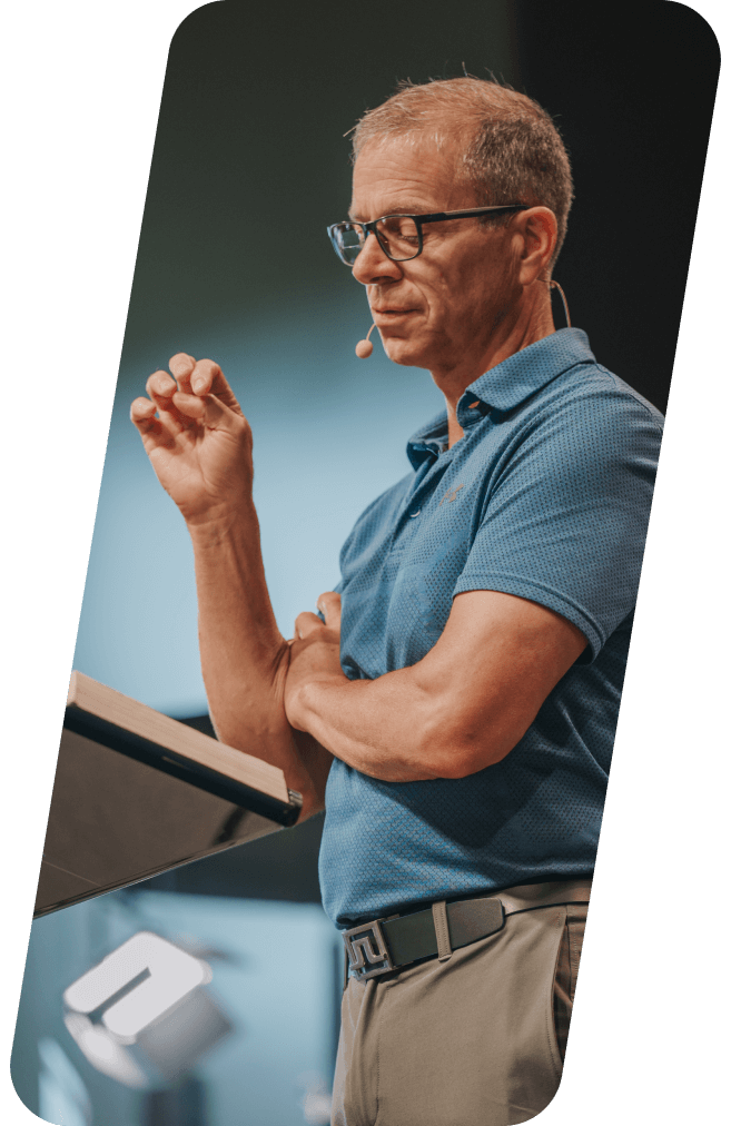 Man speaking at a podium, wearing glasses and a blue shirt, gesturing with his hand.