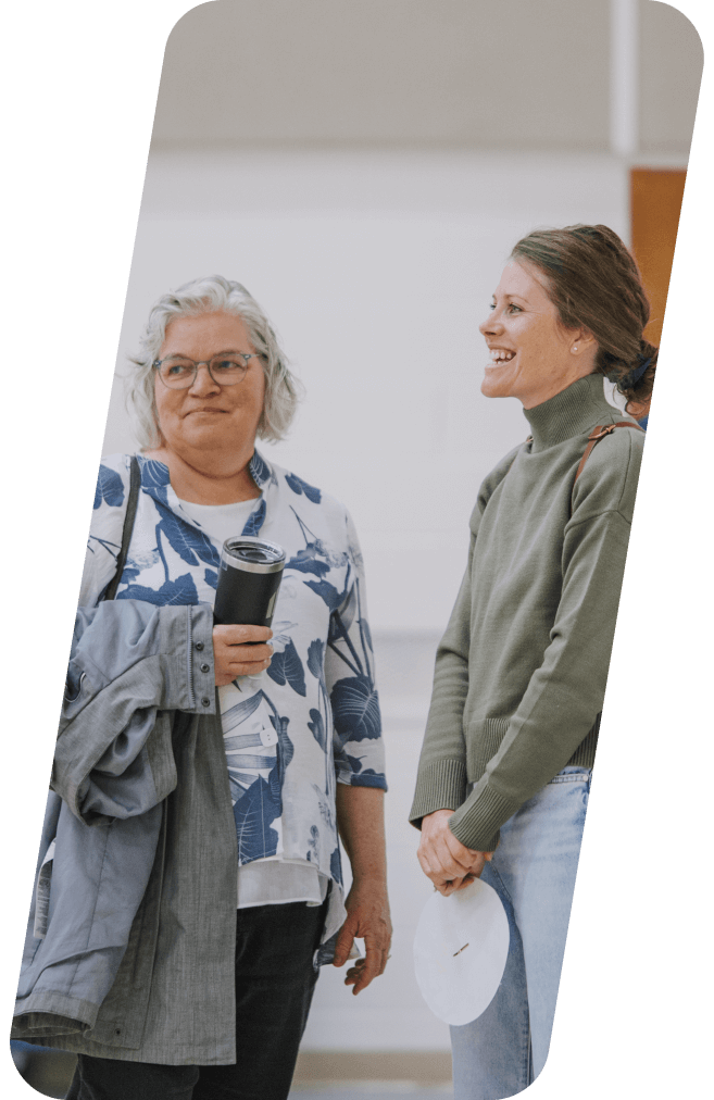 Two women chatting in a hallway, one holds a coat and coffee, the other smiles and holds a plate.