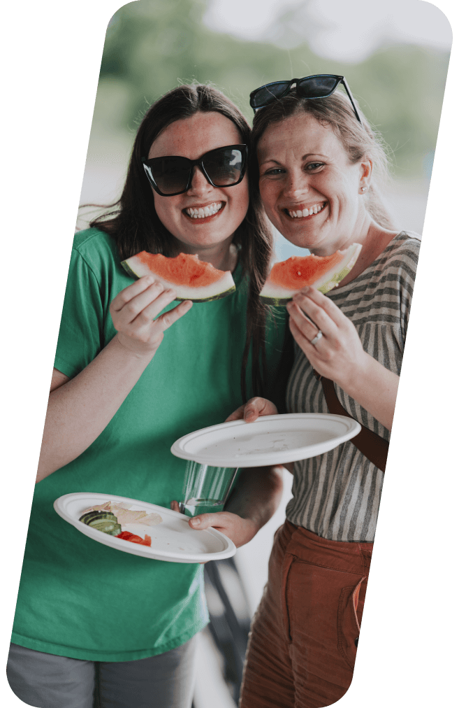 Two smiling people eating watermelon slices at an outdoor gathering.
