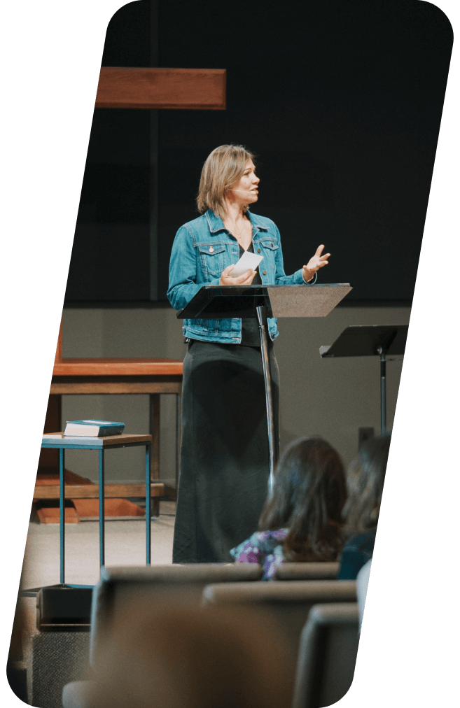 Woman speaking at a podium in an auditorium, holding papers, wearing a denim jacket and black dress.