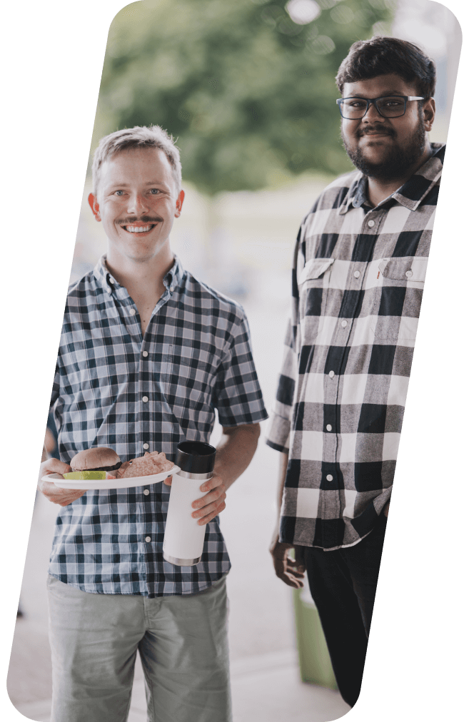 Two people with plaid shirts, one holding food and a drink, smiling outdoors.