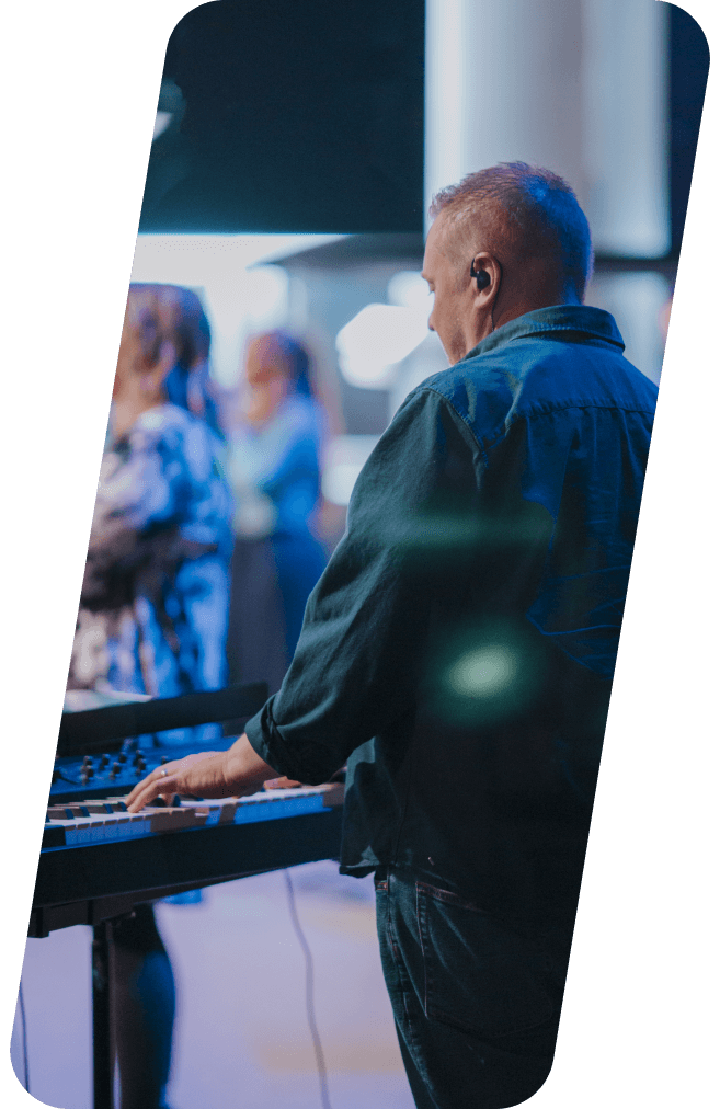 Man playing keyboard onstage, other musicians blurred in the background, blue and dim lighting.