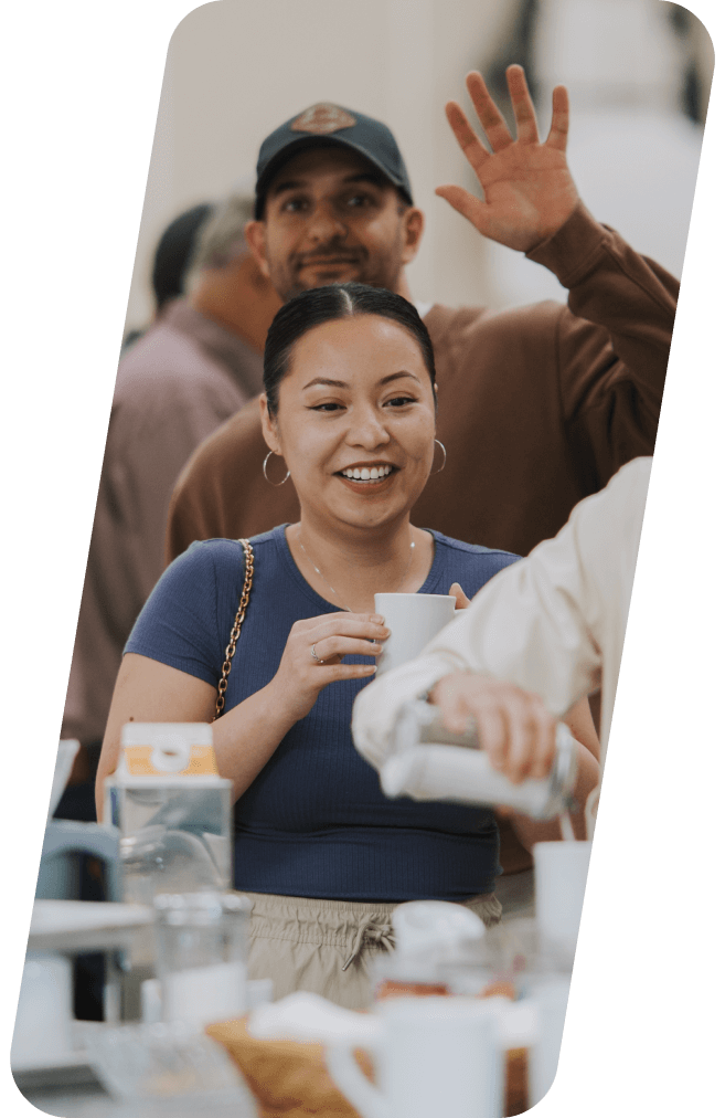Woman smiles, holding a cup. Man behind waves. Both appear at an event.