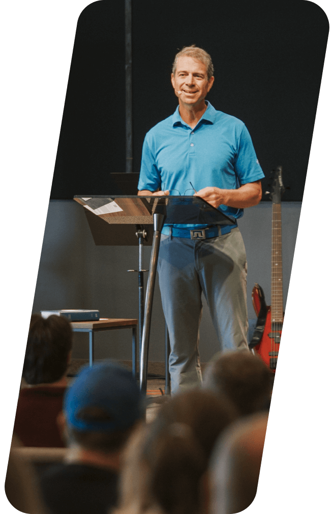 Man speaking at a podium in a church, wearing a blue shirt and jeans, facing an audience.