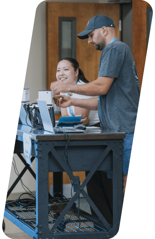 Man and woman working at a table with equipment, smiling, likely in an office setting.