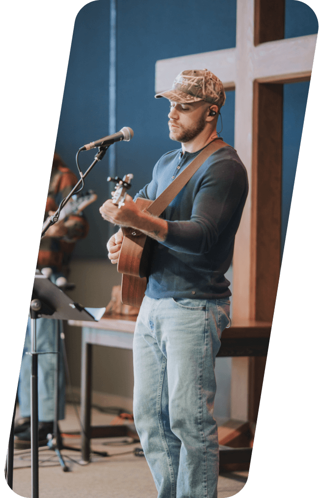 Man in camo cap plays guitar and sings at a church with a cross in the background.