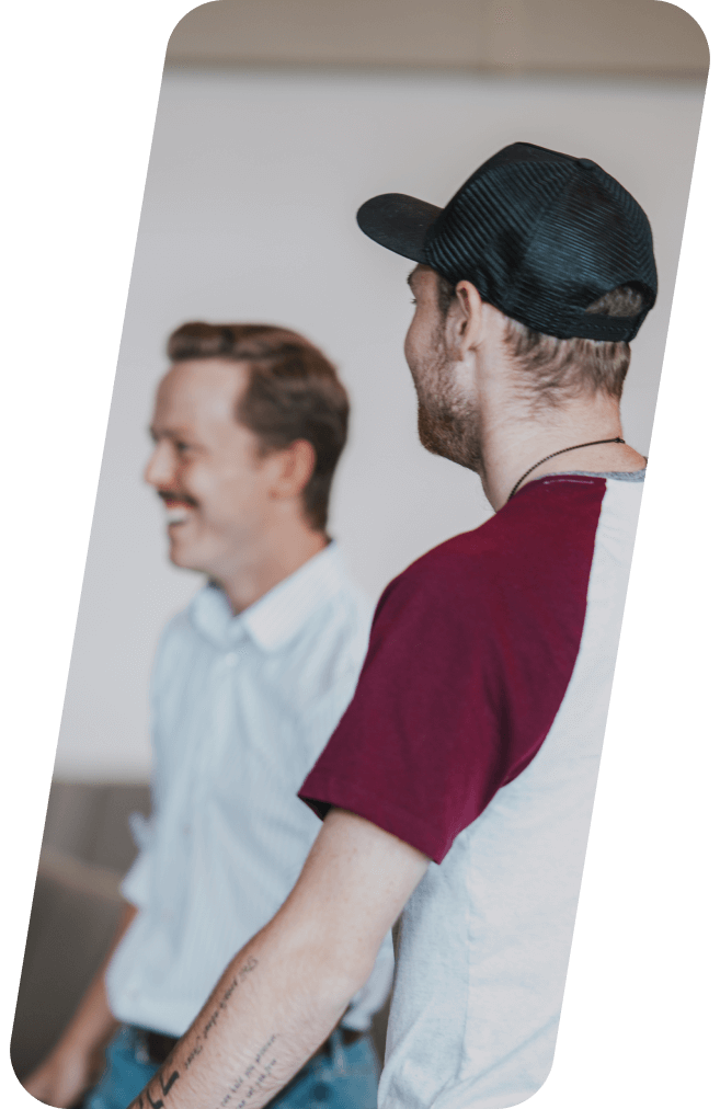 Two men smiling; one wearing a cap and burgundy shirt, the other a white shirt.