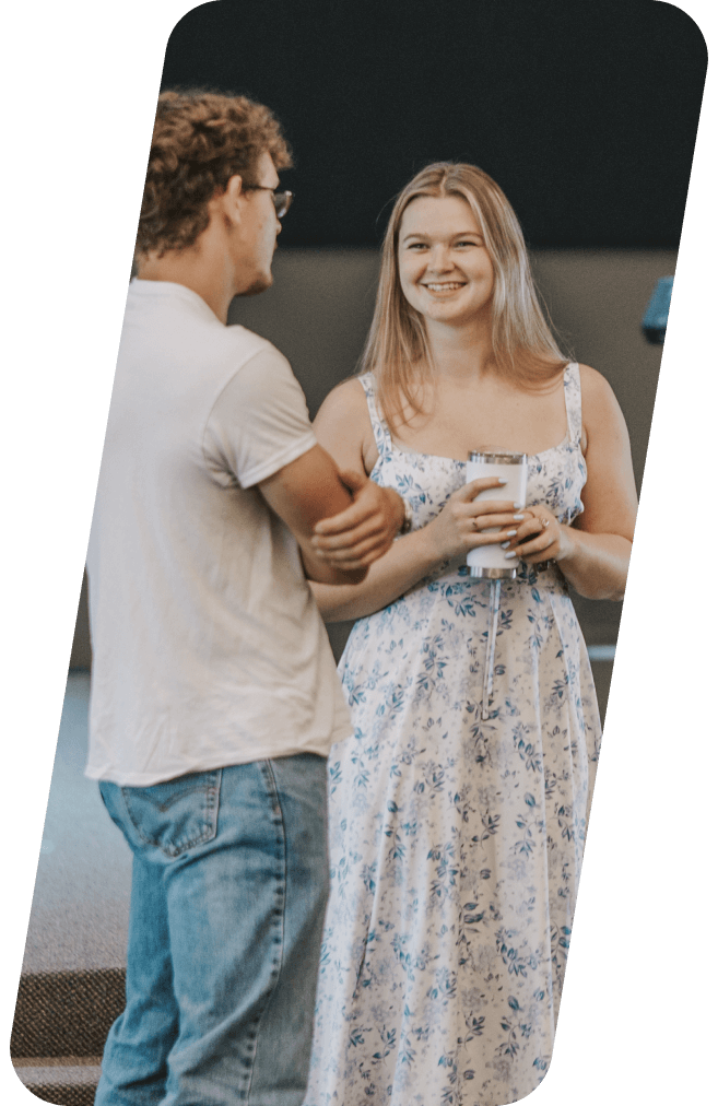 Woman in floral dress smiles, holding a cup, talking with a person wearing a white shirt and jeans.