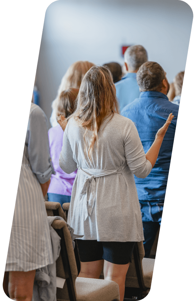 Woman with arms raised in a light-filled room, facing a crowd. Others stand nearby.