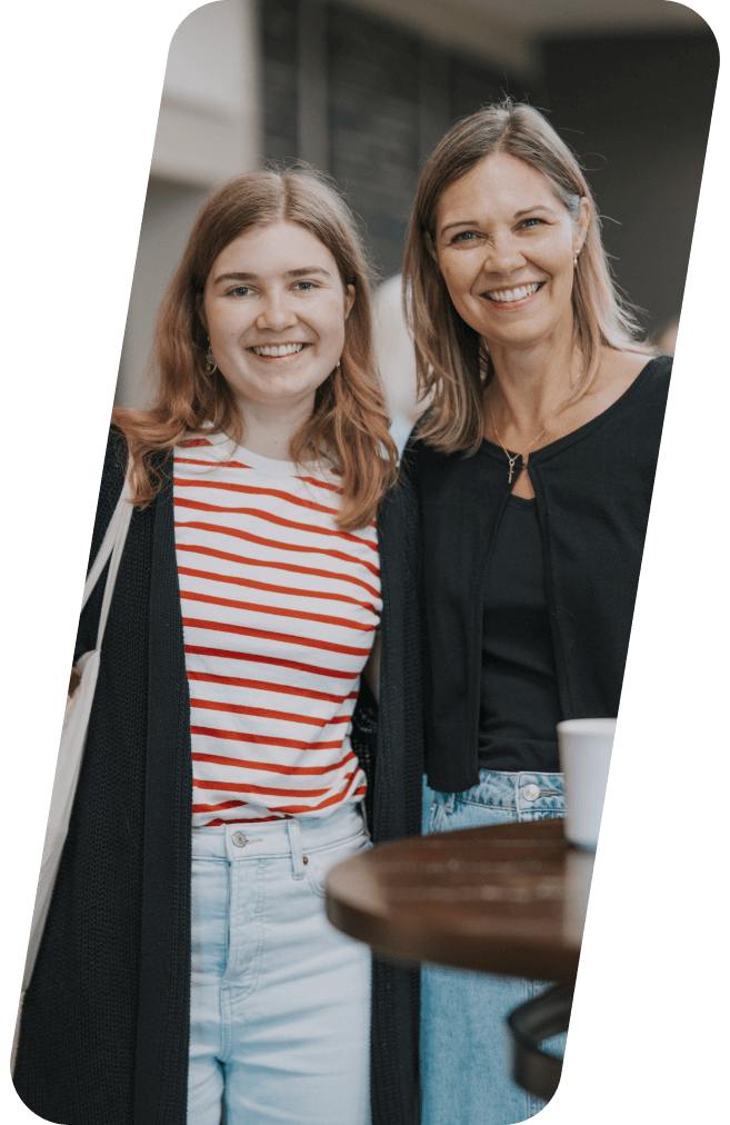 Two smiling women pose outdoors, one in a striped shirt and jeans, the other in a black top and cardigan.