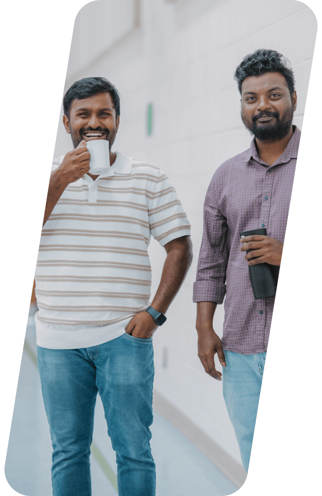 Two men smiling; one drinking from a white mug, the other holding a phone and face mask. Outdoors, casual attire.