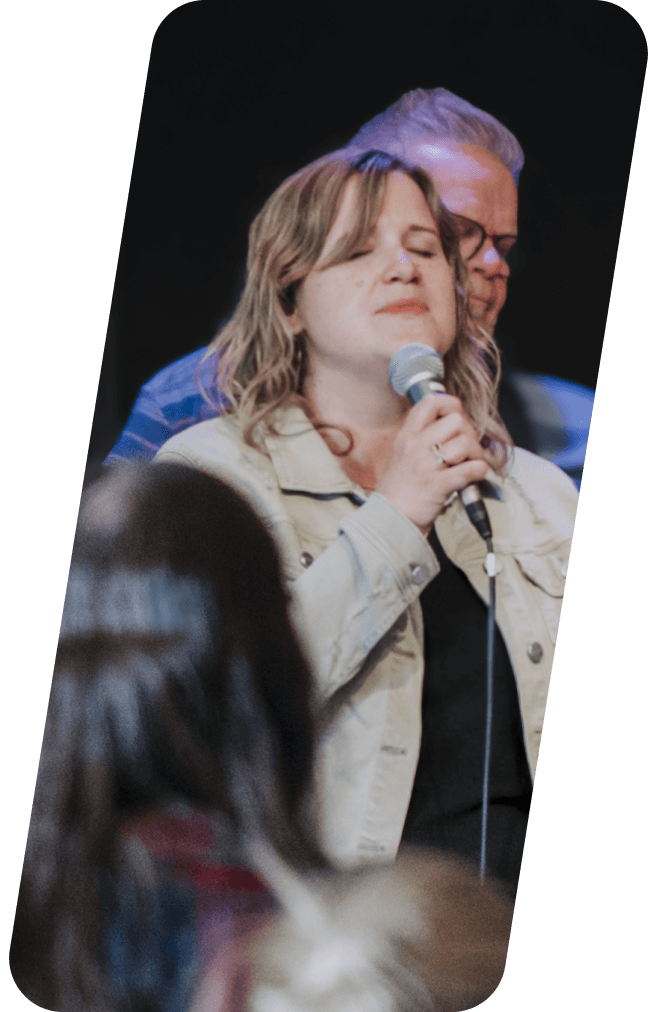 Woman singing into a microphone onstage; man's arm on her shoulder, dark background.