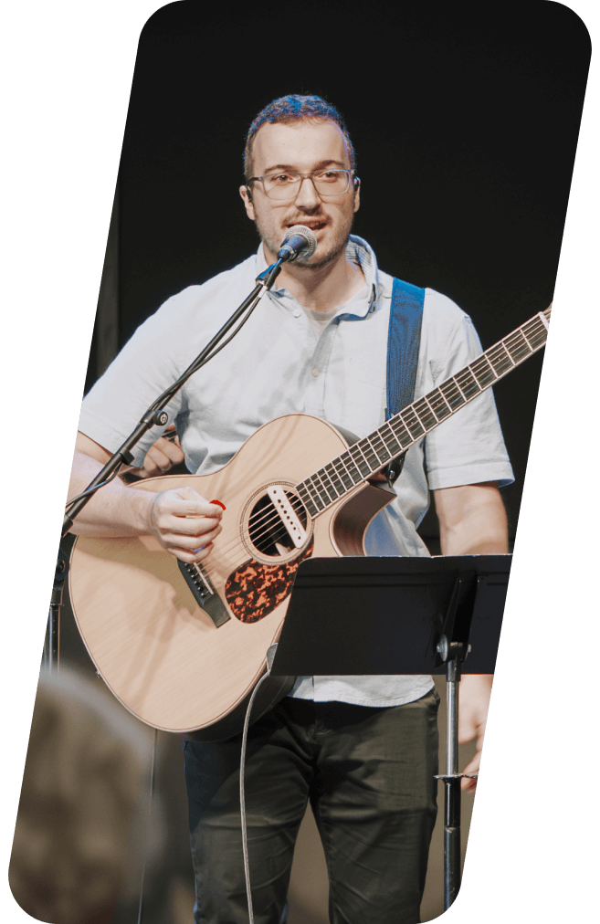 Man playing acoustic guitar and singing into a microphone onstage; black background.