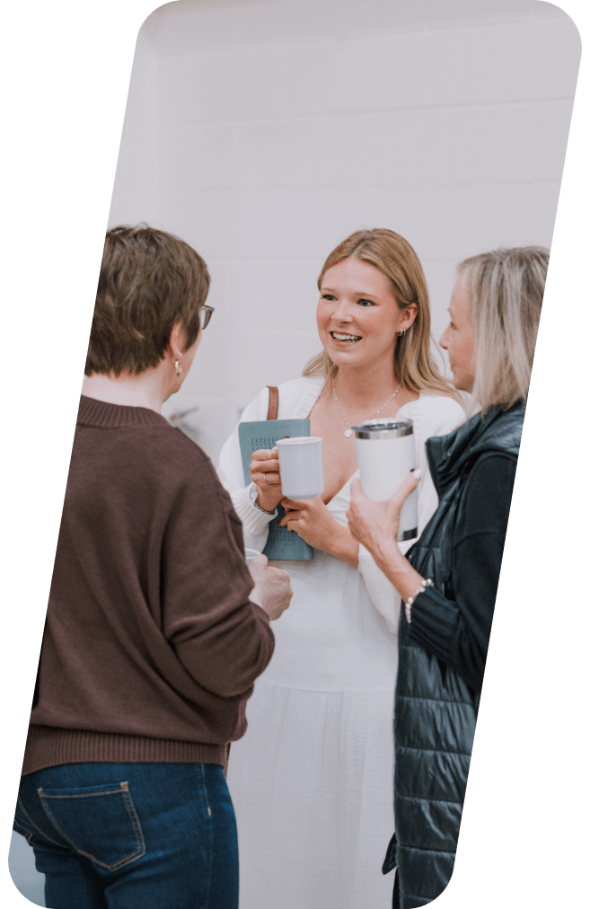 Three women smiling and chatting, holding cups, near a white wall.