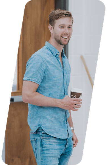 Man in blue shirt and jeans, holding a coffee cup, smiling. Standing near a wooden door.