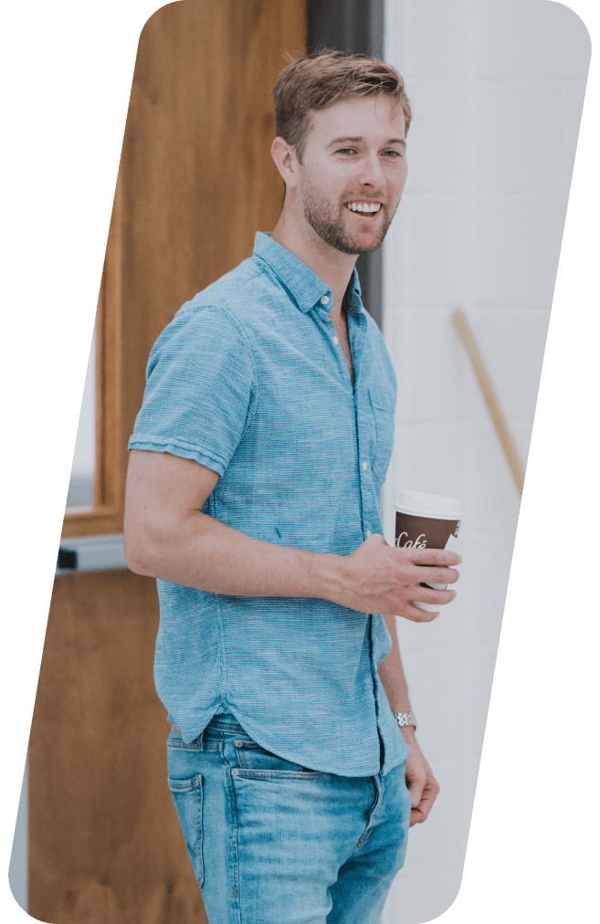 Man in blue shirt and jeans, holding a coffee cup, smiling. Standing near a wooden door.
