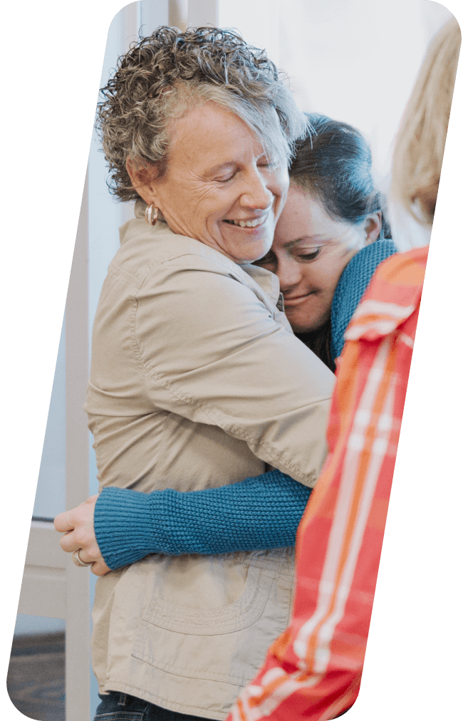 Woman hugging another woman, both smiling; light-colored clothing, warm expression.