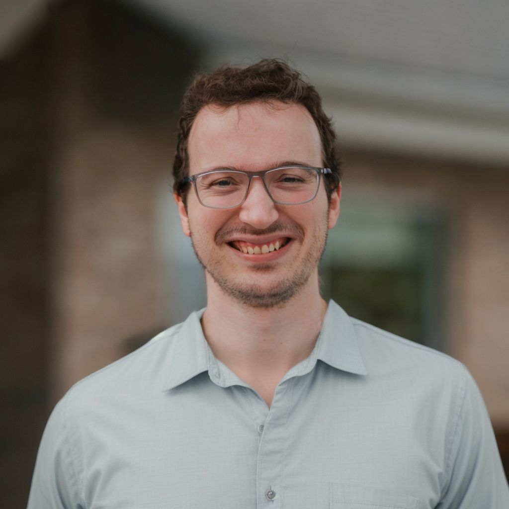 Smiling person wearing glasses and a light blue shirt, outdoors.