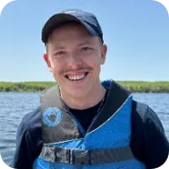 Man wearing a blue life vest and hat smiles while on a boat, against a lake.