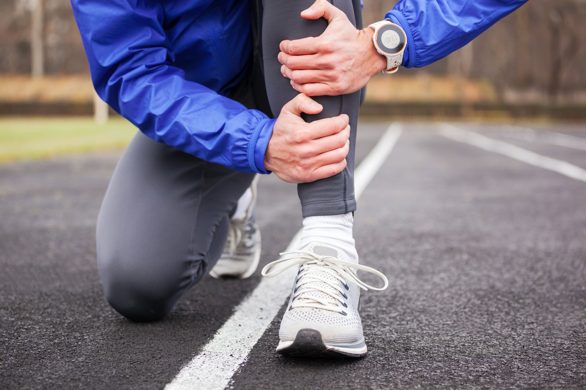 Athlete kneeling on a track, clutching lower leg in pain, wearing athletic clothing and a smartwatch.