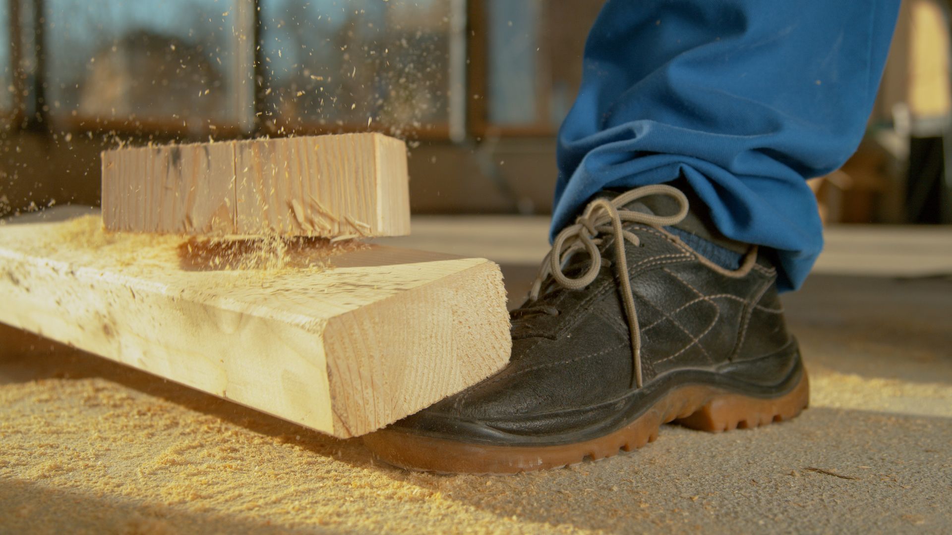 Person in blue work pants and safety boots near cut wood with sawdust.