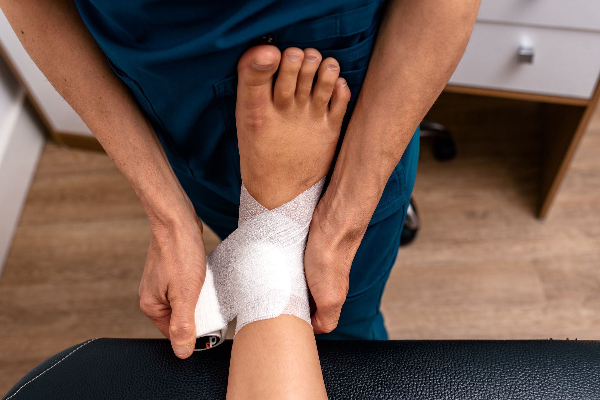 Person wrapping a bandage around another person’s ankle. Indoor, clinical setting.