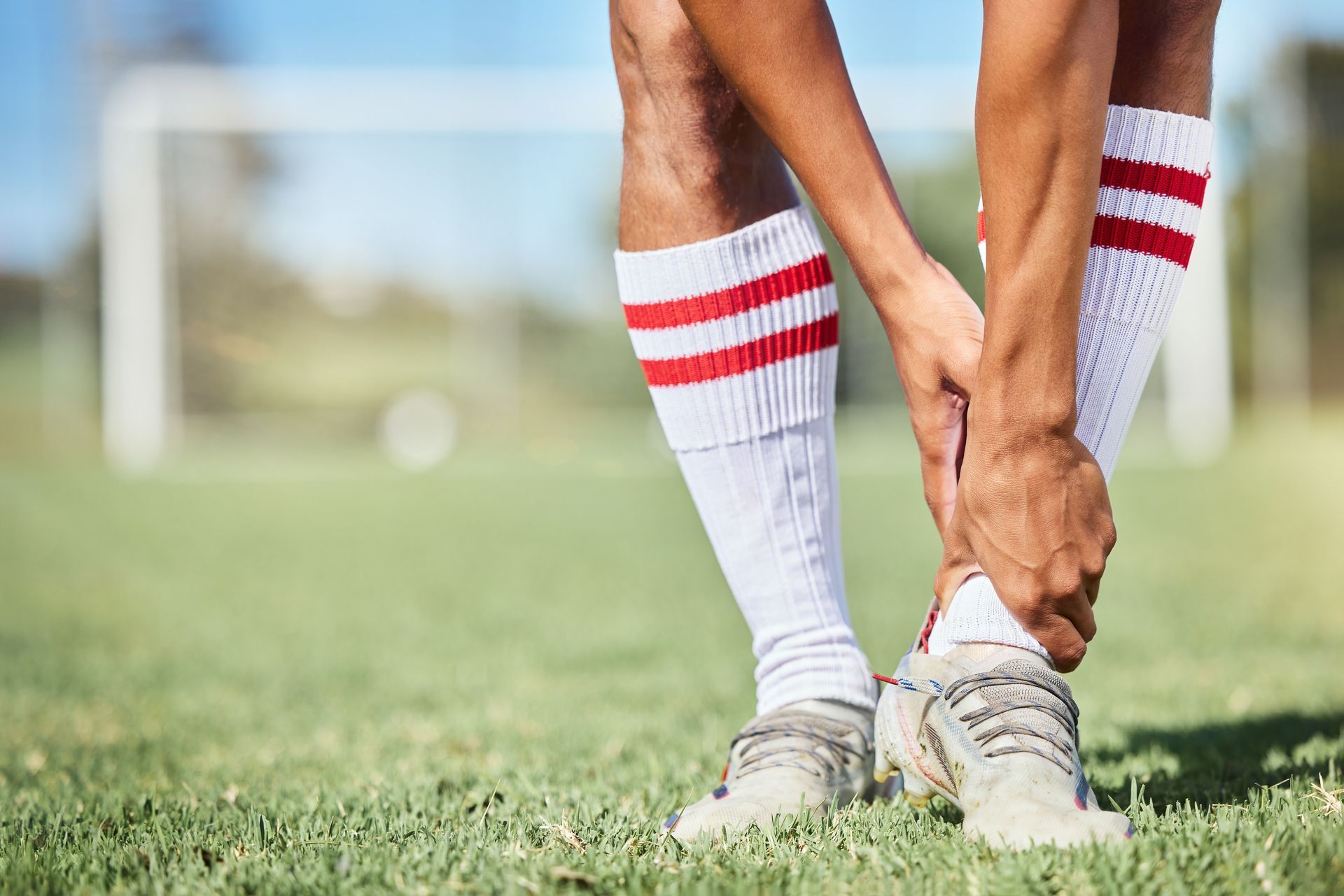 Soccer player holding injured ankle on a grassy field, wearing white socks with red stripes.