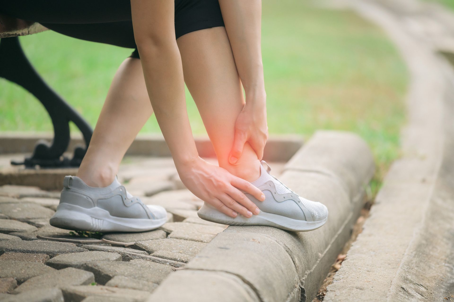 Person holding ankle, appears to be in pain, sitting on a bench in a park.