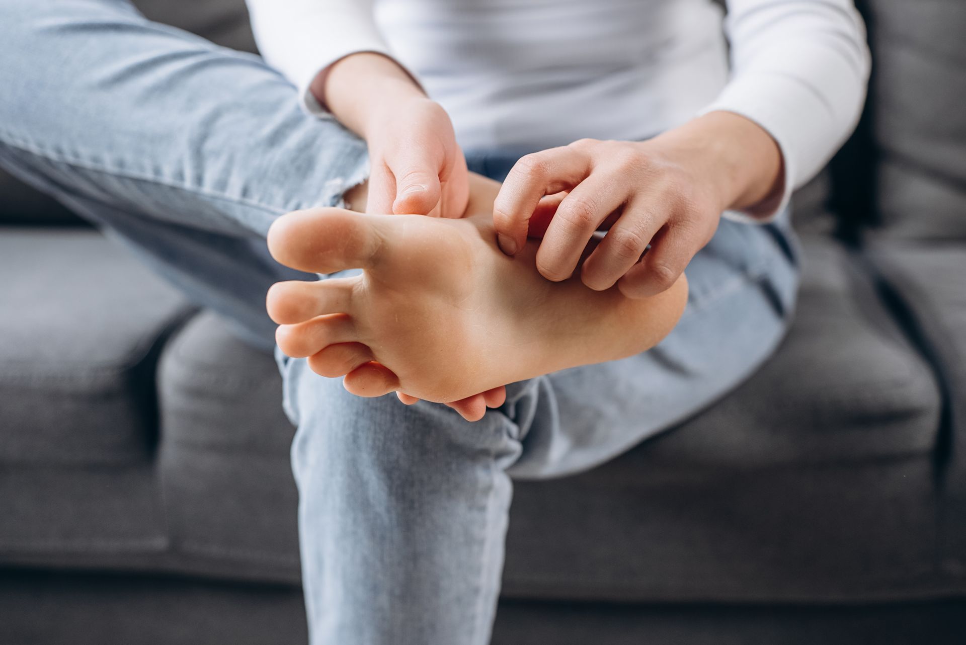 Person scratching foot, sitting on a gray couch, wearing jeans and a white shirt.