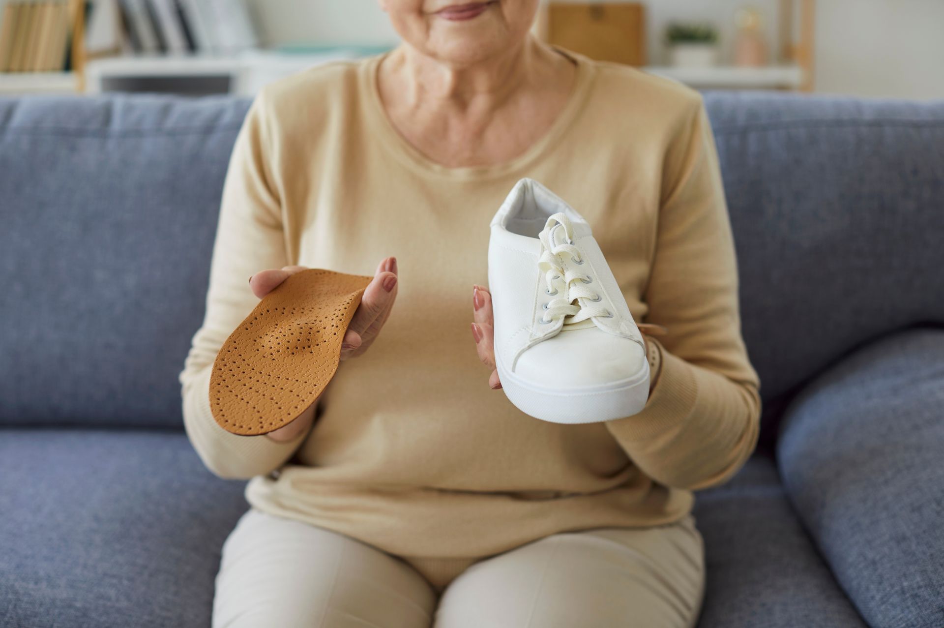 Woman holding a shoe and orthopedic insole, sitting on a blue couch. Indoors.