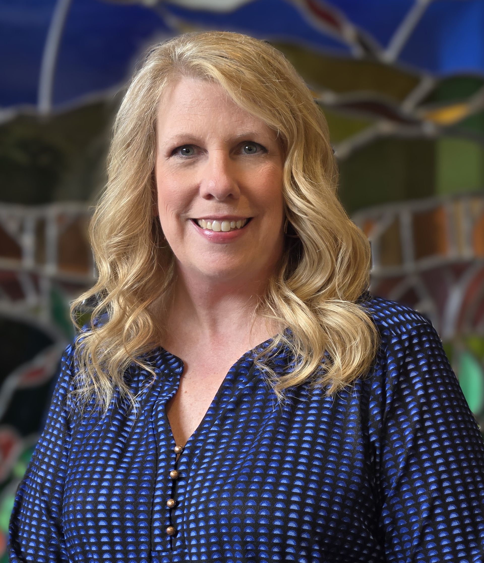 Woman with blonde hair smiles, wearing a black jacket, standing next to a brick wall.
