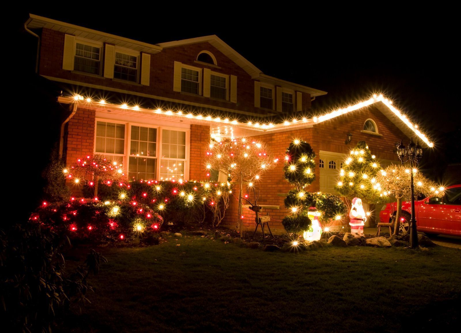 A brick house decorated with bright Christmas lights at night, with a red car parked nearby.