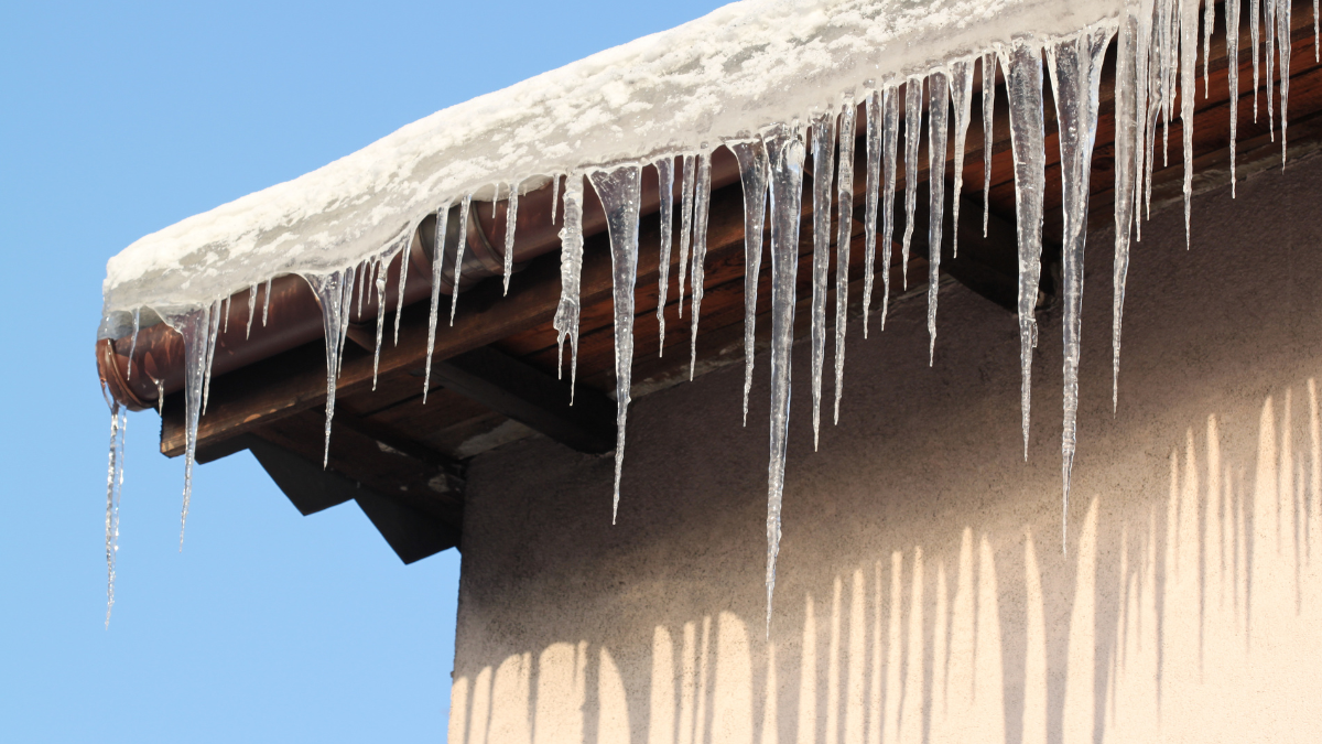 Icicles hanging from a roof's edge, reflecting sunlight against a beige wall and blue sky.
